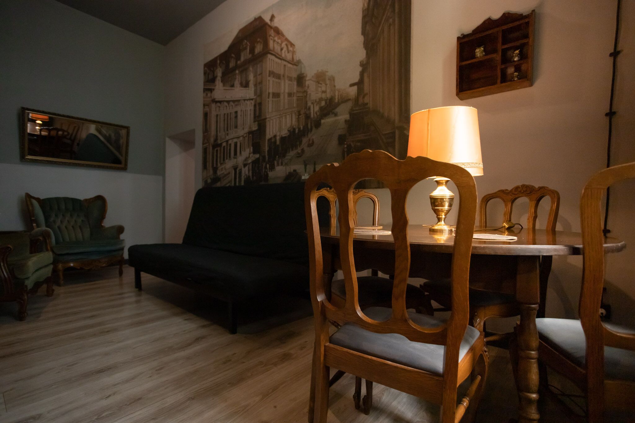 A living room with a wooden floor, a green armchair, a black sofa, a dining table with chairs, a lamp, and a cityscape mural on the wall.