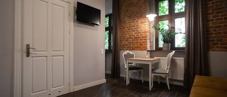 A room with a white door, a TV mounted on the wall, a dining table with chairs, a window with a view of greenery, and a brick wall.