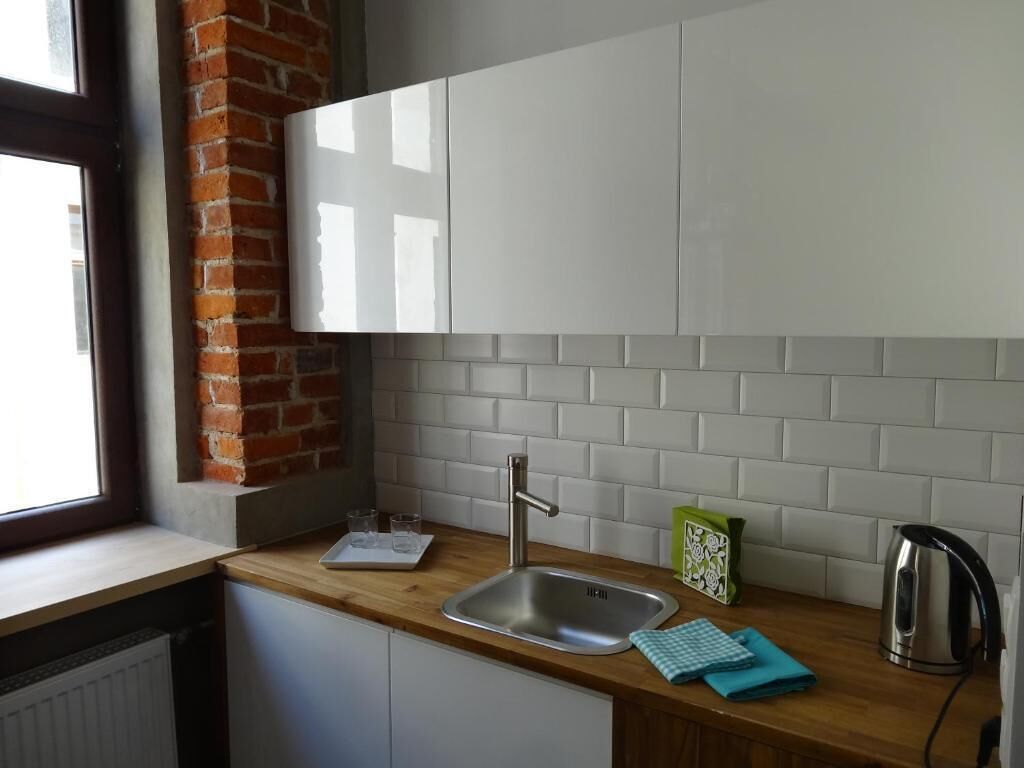 Una cocina con ventana, armarios blancos, un lavamanos de acero inoxidable, un hervidor y una pared de ladrillo.