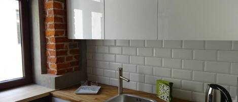 Una cocina con ventana, armarios blancos, un lavamanos de acero inoxidable, un hervidor y una pared de ladrillo.