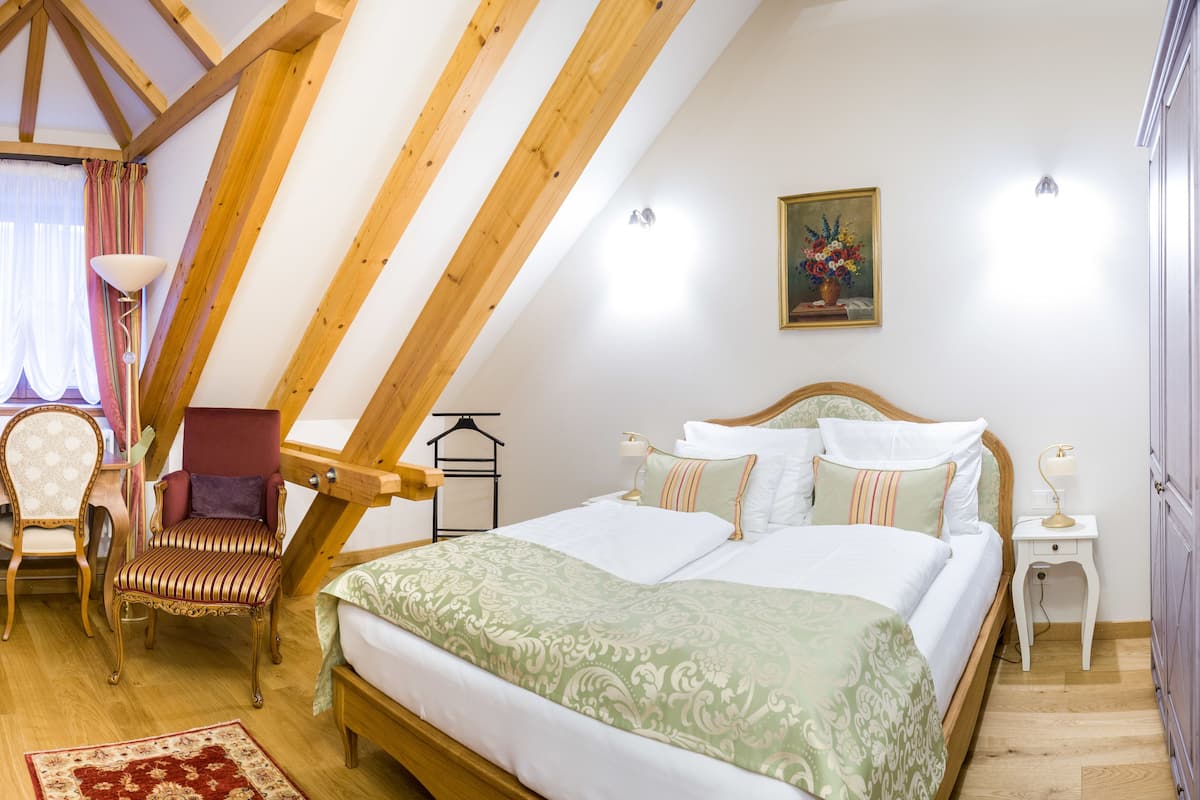 Une chambre avec un plafond en pente, un lit recouvert d’une couvre-lit à motifs verts, une chaise en bois, un bureau avec une chaise et une armoire.