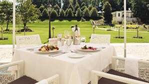 Un área de comedor al aire libre con una mesa preparada para dos personas, rodeada de sillas de mimbre blanco, con vistas a un jardín cuidado y un edificio al fondo.