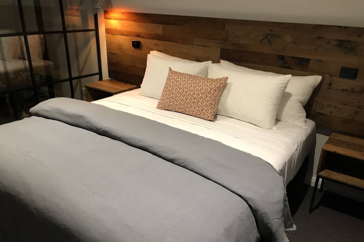 Uma cama bem arrumada com um cobertor cinza e duas almofadas, uma cabeceira de madeira e uma mesa de cabeceira com um abajur.