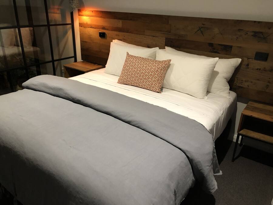 A neatly made bed with a grey blanket and two pillows, a wooden headboard, and a bedside table with a lamp.