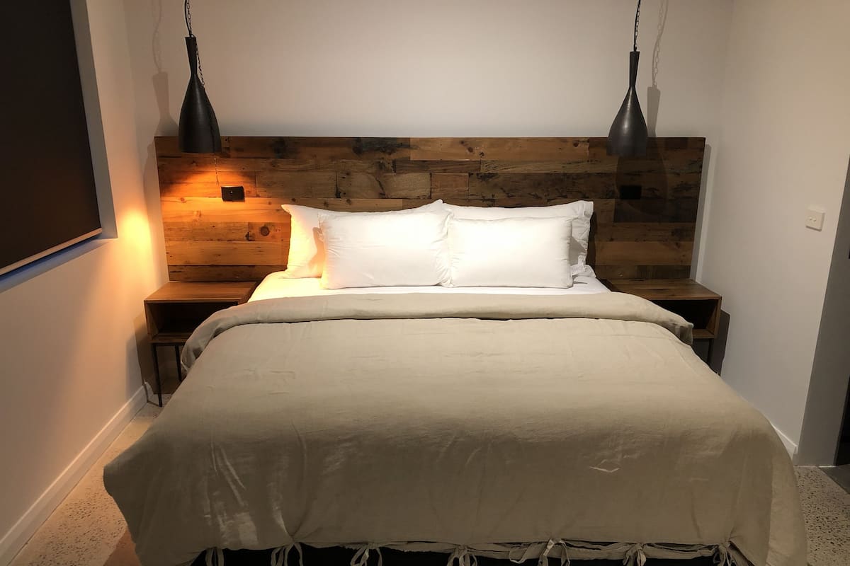 Cama com cabeceira de madeira, almofadas brancas e manta bege. Há duas mesas de cabeceira com abajures.