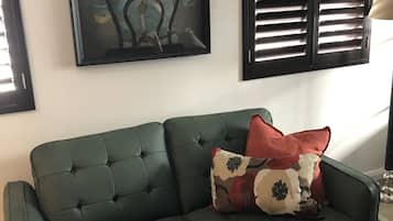 A green sofa with a patterned pillow, a framed painting on the wall, and a window with blinds.
