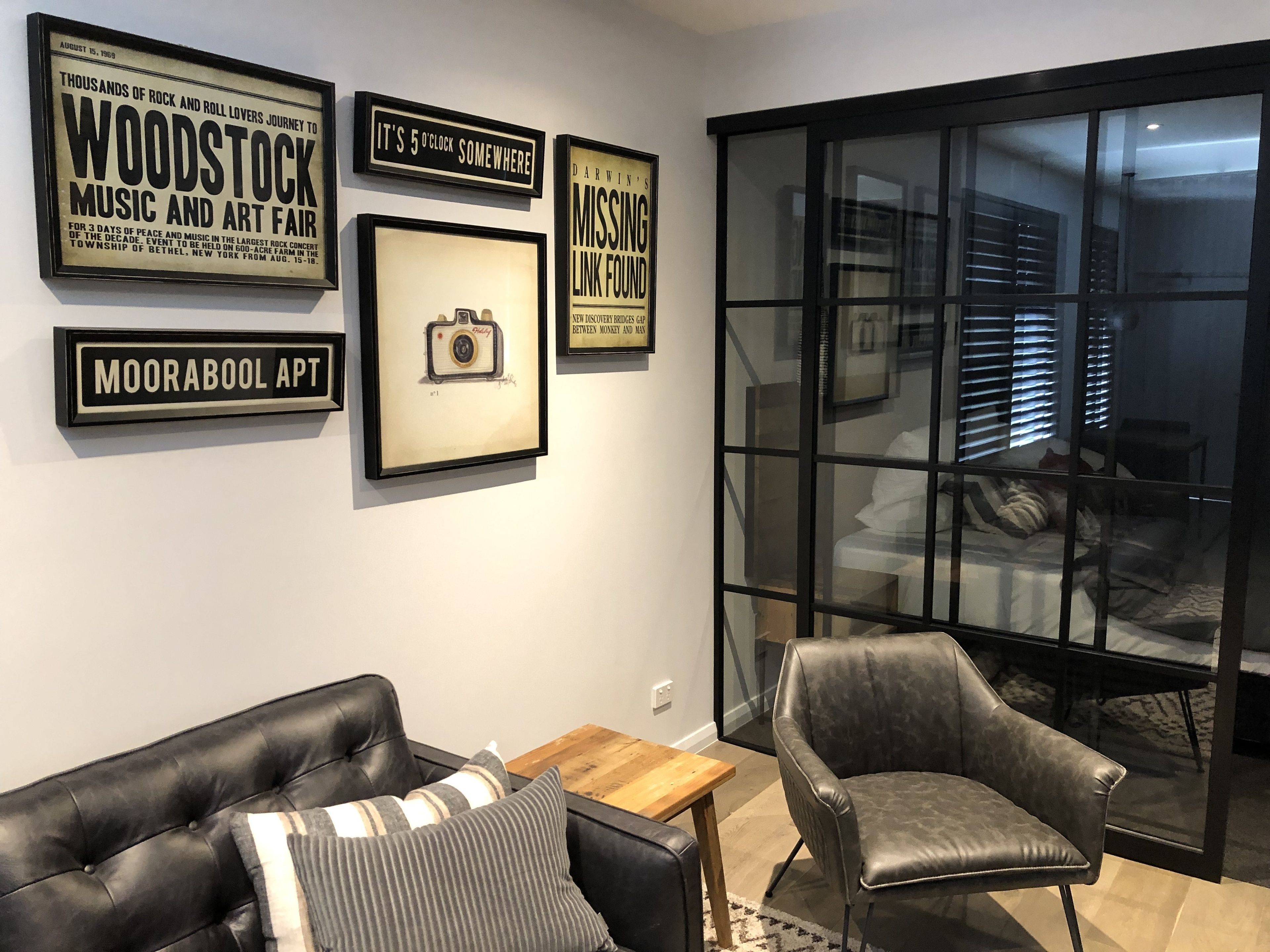 A modern living room with a black leather sofa, a grey armchair, and a wooden coffee table. There are framed posters on the wall, including one that says "WOODSTOCK MUSIC AND ART FAIR".