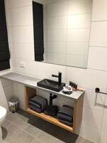Un baño moderno con encimera gris, lavabo negro y estante de madera con toallas.