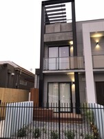 Un edificio moderno de dos pisos con balcón, ventanas grandes y entrada protegida por un portón.