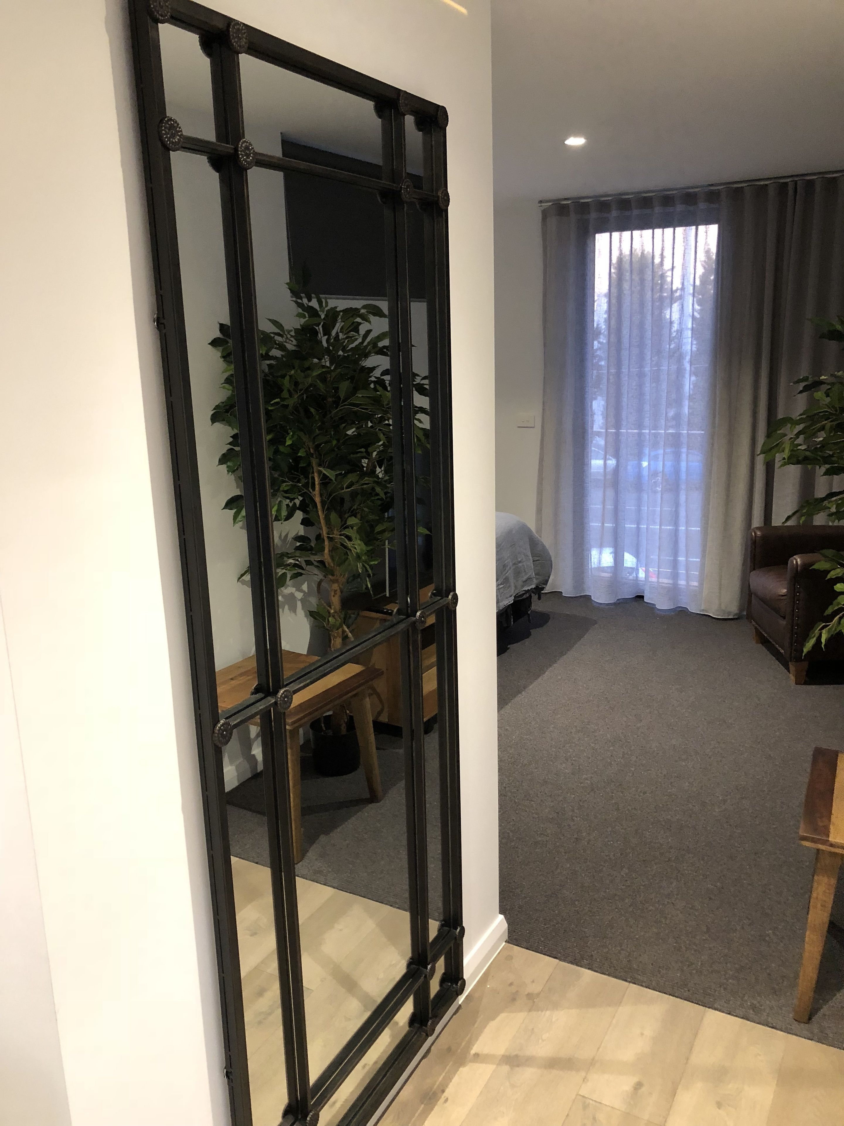 A room with a large mirror, a wooden floor, a potted plant, a sofa, and a window with curtains.