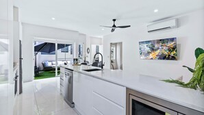 Eine moderne Küche mit weißen Schränken, einem eingebauten Geschirrspüler und einer großen Kochinsel mit Spüle. Ein Deckenventilator, eine Klimaanlage und ein an der Wand befestigtes Fernsehgerät zeigen eine Stadtansicht.