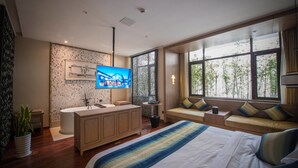 Una habitación de hotel moderna con una cama grande, un televisor montado en la pared, un tocador de madera y un asiento junto a la ventana con vistas a árboles.