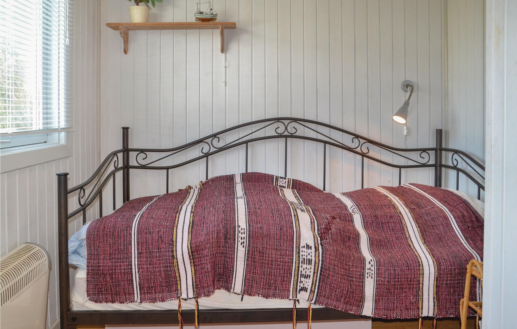 Ein Bett mit Metallgestell, eine rot-weiße Decke, eine kleine Ablage mit einer Pflanze und einem Spielschiff sowie eine wandmontierte Leuchte.