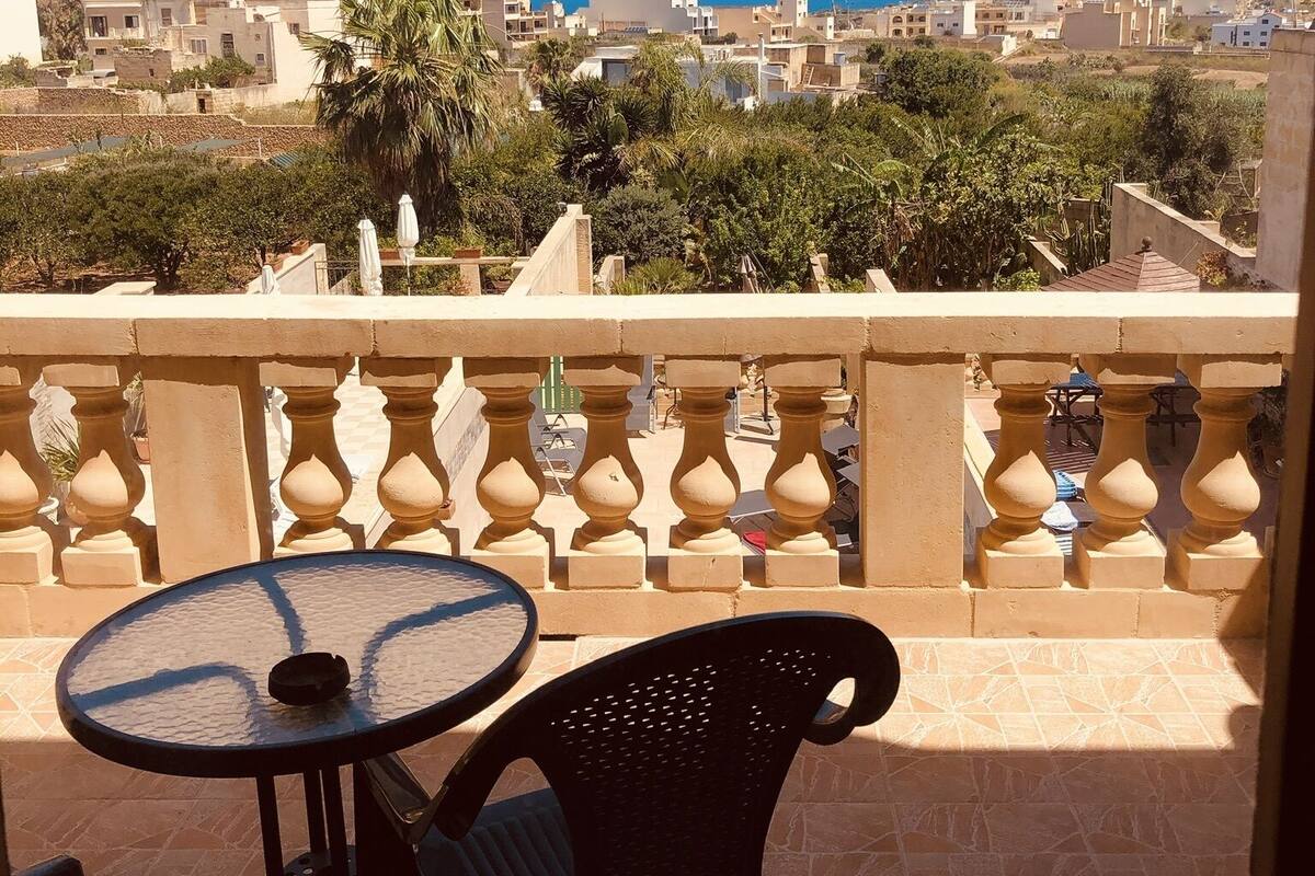 A balcony with a table and chairs, overlooking a coastal town with buildings and palm trees.