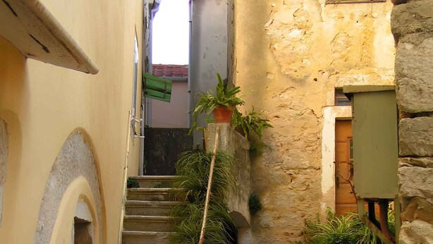 Un stretto vicolo con gradini in pietra che conducono a una porta, affiancato da edifici con finestre ad arco e piante in vaso.