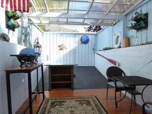 A covered patio with a wooden table and chairs, a small shelf, and hanging planters. - PORTHOLE: Private, Peaceful with a locked patio for relaxing and bikes. (Port Angeles)