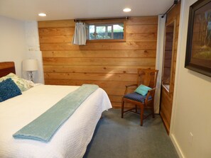 A bedroom with a wooden wall, a bed with a blue blanket, a chair with a blue cushion, and a window with a curtain. - PORTHOLE: Private, Peaceful with a locked patio for relaxing and bikes. (Port Angeles)