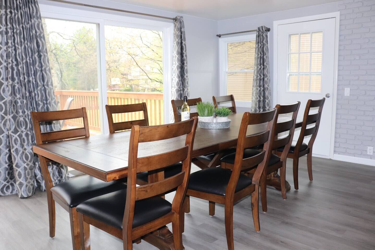 A dining area with a wooden table and chairs, large windows with curtains, and a brick wall.