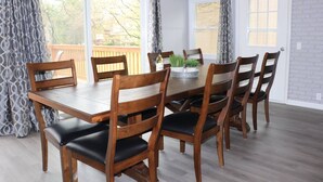 A dining area with a wooden table and chairs, large windows with curtains, and a brick wall.