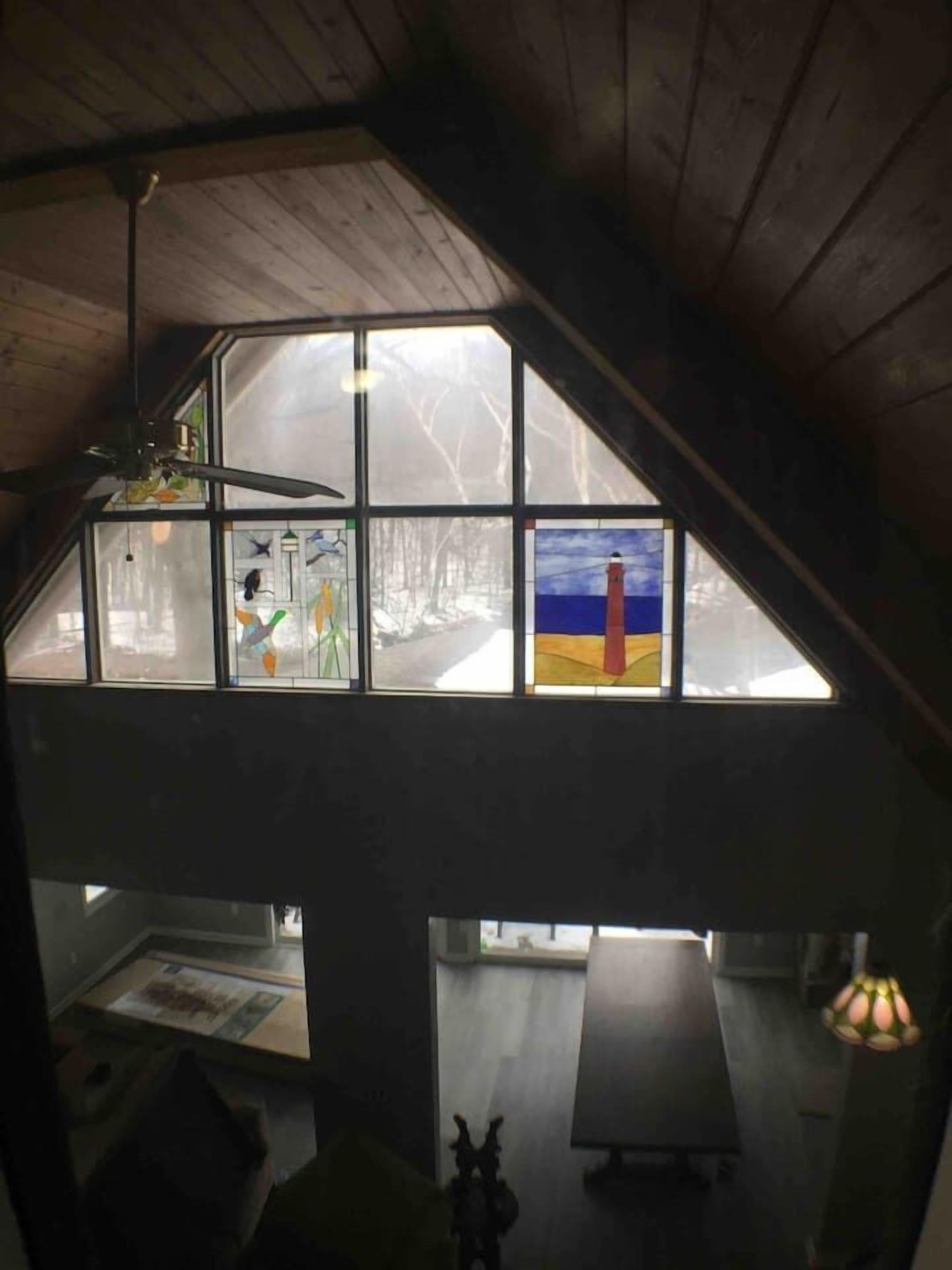 Une pièce de style loft avec un plafond en pente, de grandes fenêtres ornées d’œuvres d’art, un ventilateur de plafond et une lampe de table.