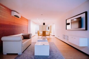 Una sala de estar moderna con un sofá, una mesa de centro, un televisor montado en la pared y un área de comedor al fondo.