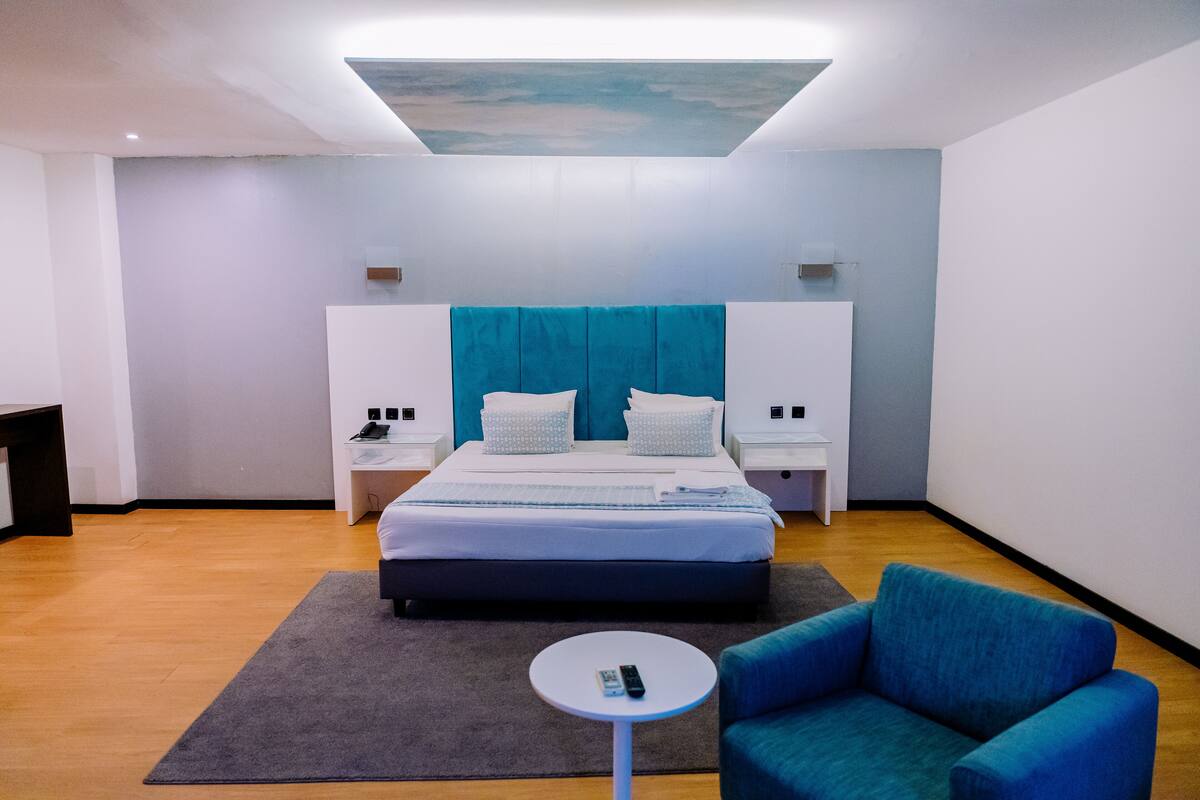 A modern hotel room with a large bed, two bedside tables, a small round table, and a blue armchair.