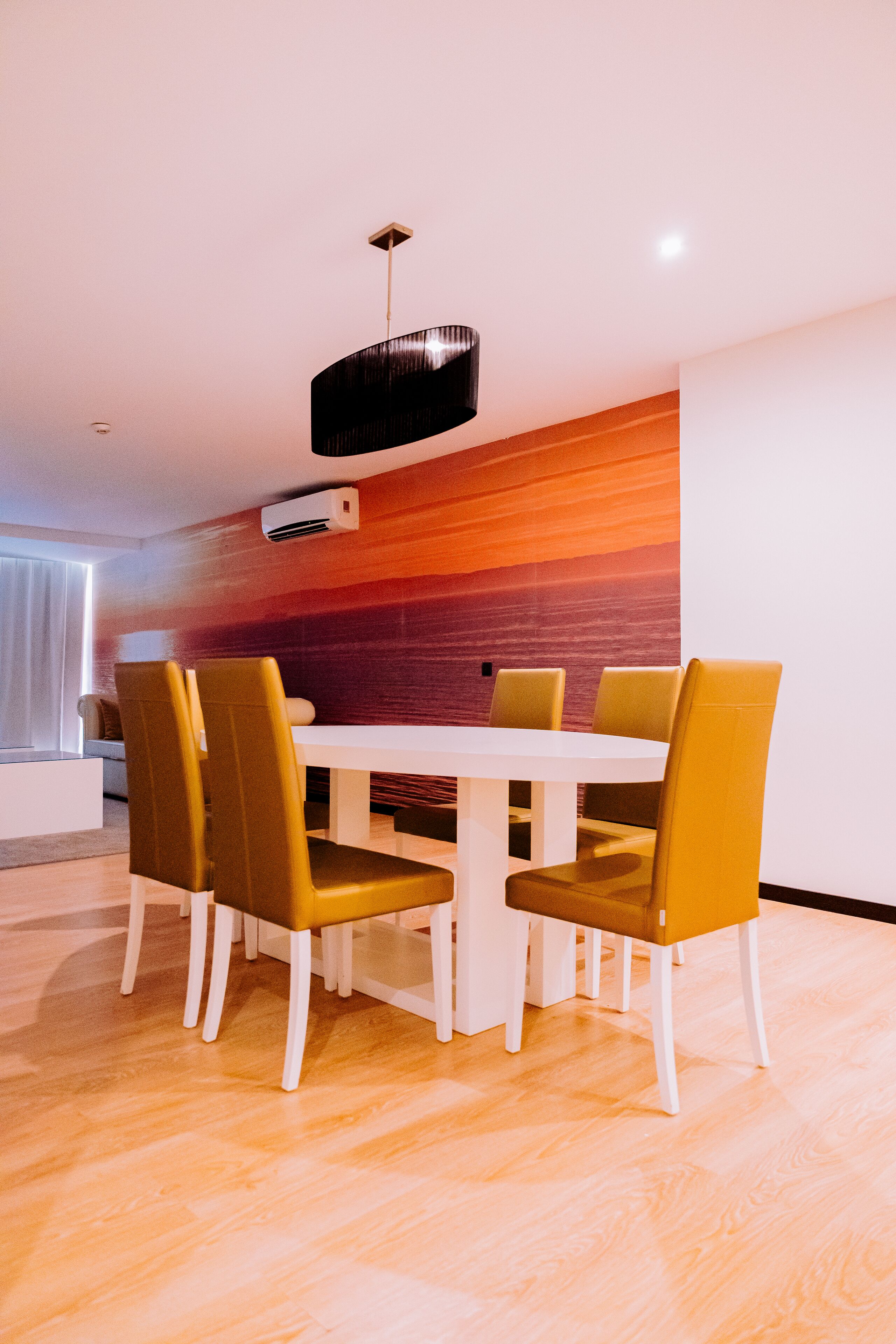 Un comedor moderno con una mesa blanca y cuatro sillas, una lámpara colgante negra y un equipo de aire acondicionado en la pared.