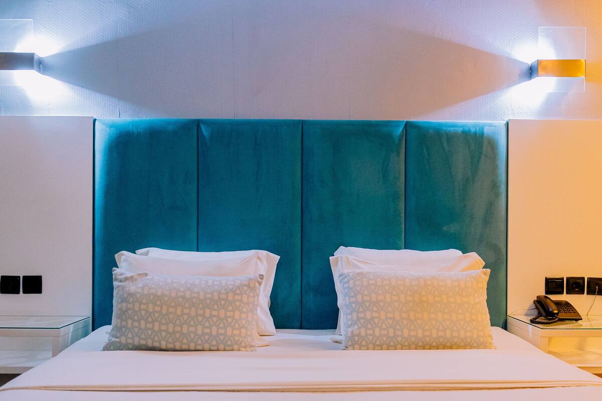 A hotel room with a large bed, two patterned pillows, a teal headboard, and bedside lamps.
