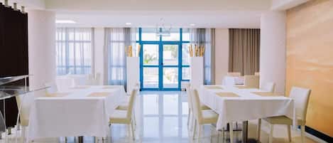 A hotel dining room with tables set for service, a clear view of the outdoors through large windows, and a modern interior design.