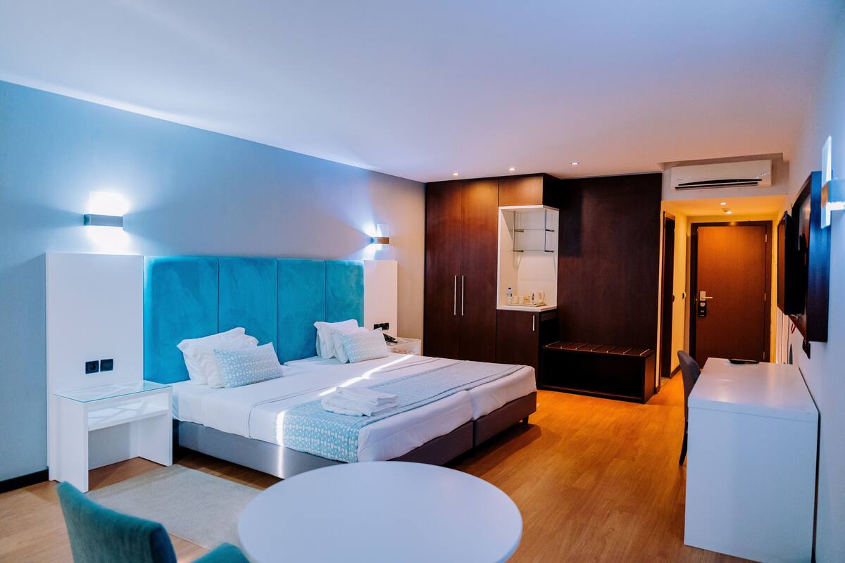 A modern hotel room with a bed, bedside tables, a desk, a round table, and a TV.