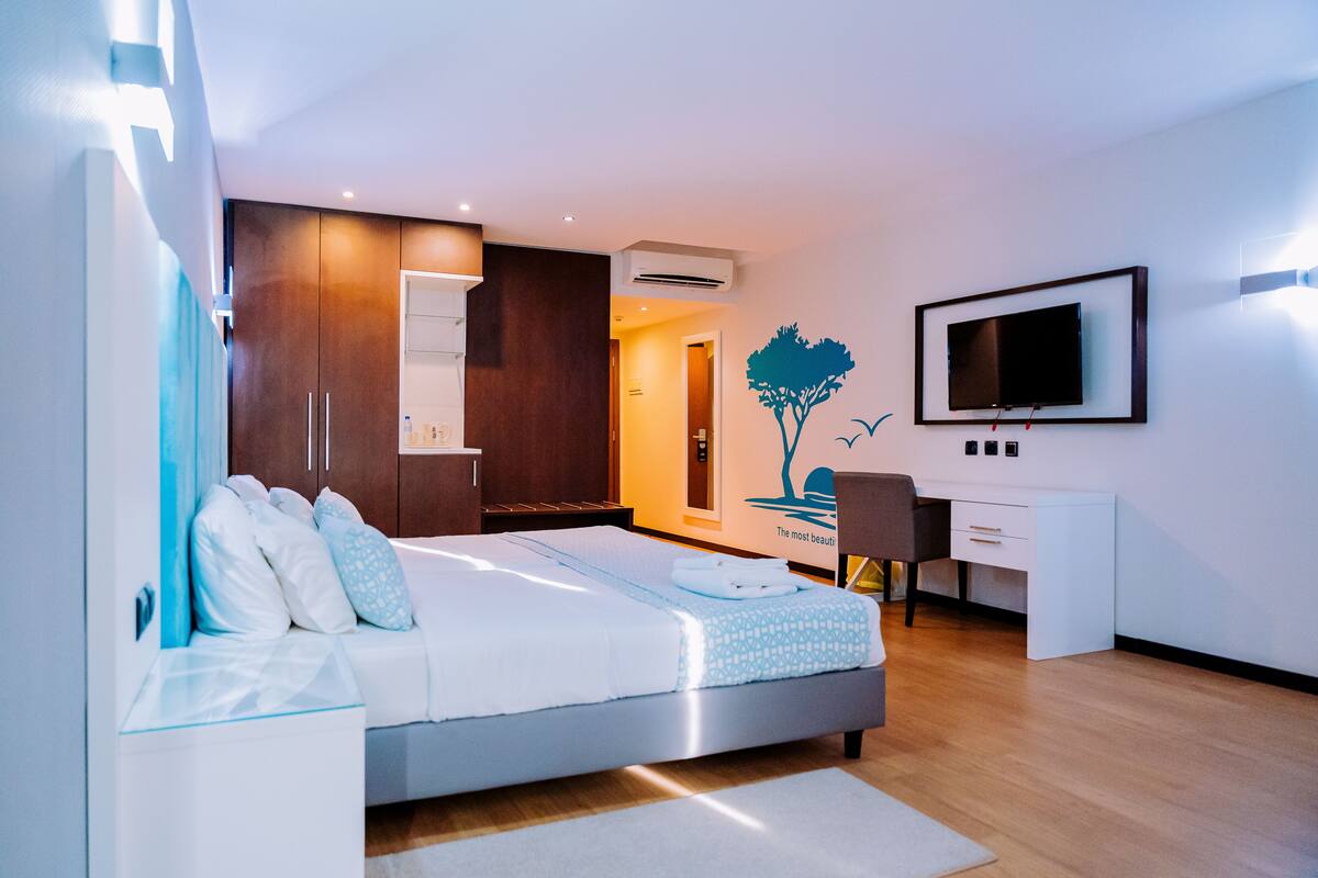 A modern hotel room with a bed, a desk, a chair, a TV, and an air conditioning unit.