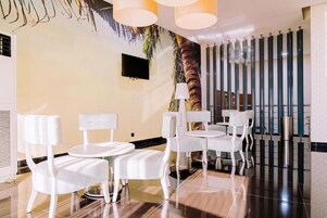 Un moderno vestíbulo de hotel con sillas y mesas blancas, un televisor de pantalla plana, un mural de palmera y una pared decorativa con elementos verticales.