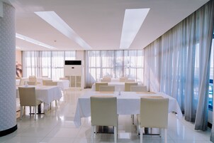 Un comedor amplio con varias mesas y sillas, ventanas grandes con cortinas ligeras y un diseño moderno de techo.