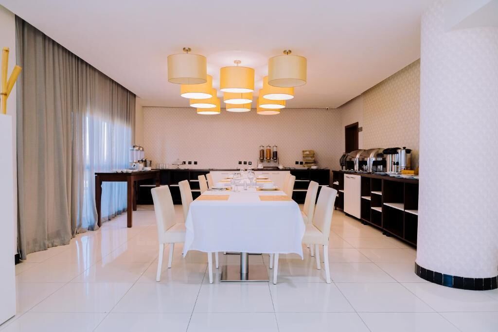 Ein moderner Essbereich mit einem weißen Tisch, gedeckt für vier Personen, umgeben von Stühlen. Über dem Tisch befinden sich Pendelleuchten, und zur Rechten ist eine Küche mit Geräten und einem Kühlschrank zu sehen.