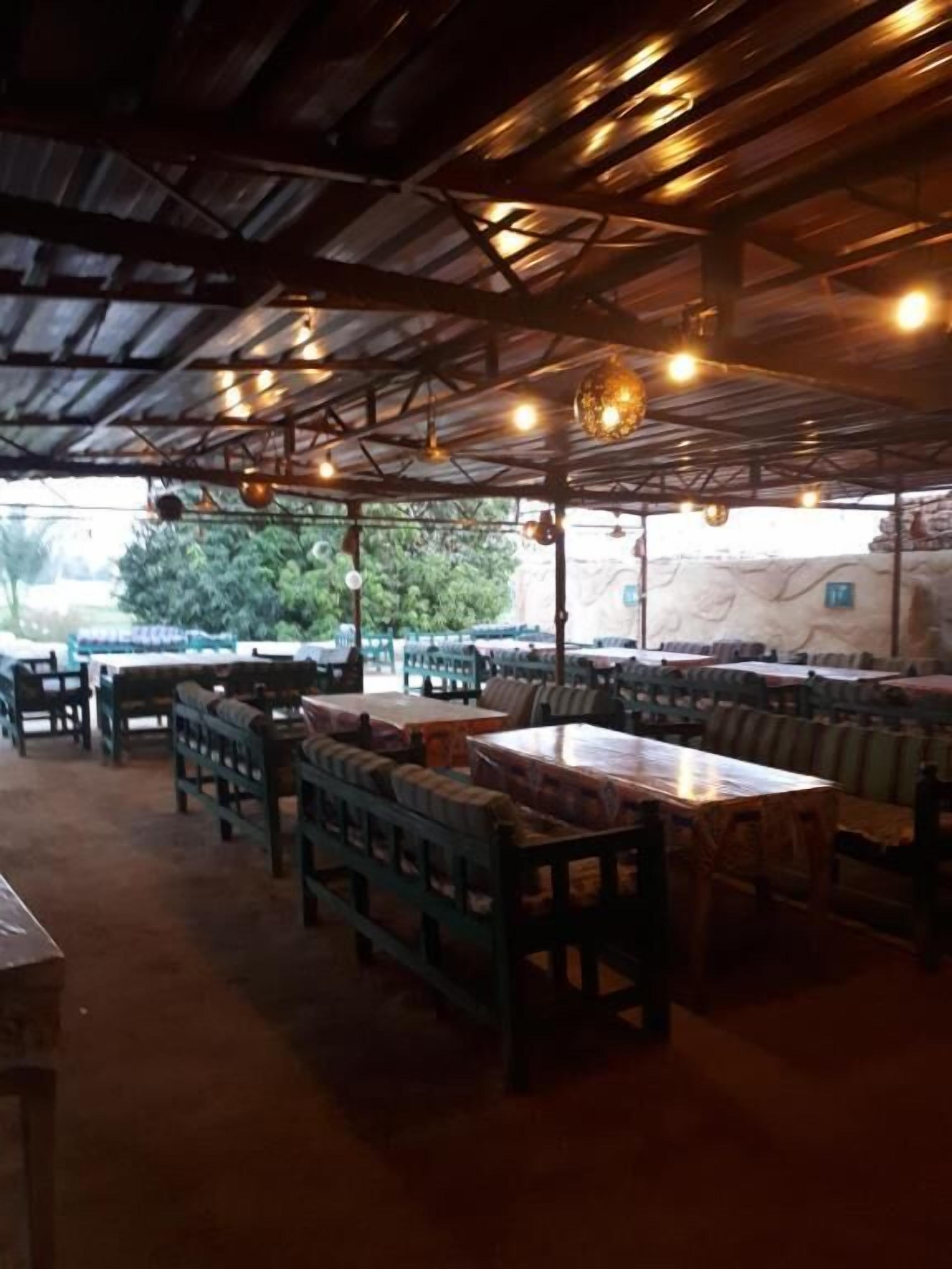 Restaurante ao ar livre com bancos e mesas de madeira, sob uma cobertura de metal com luminárias pendentes.