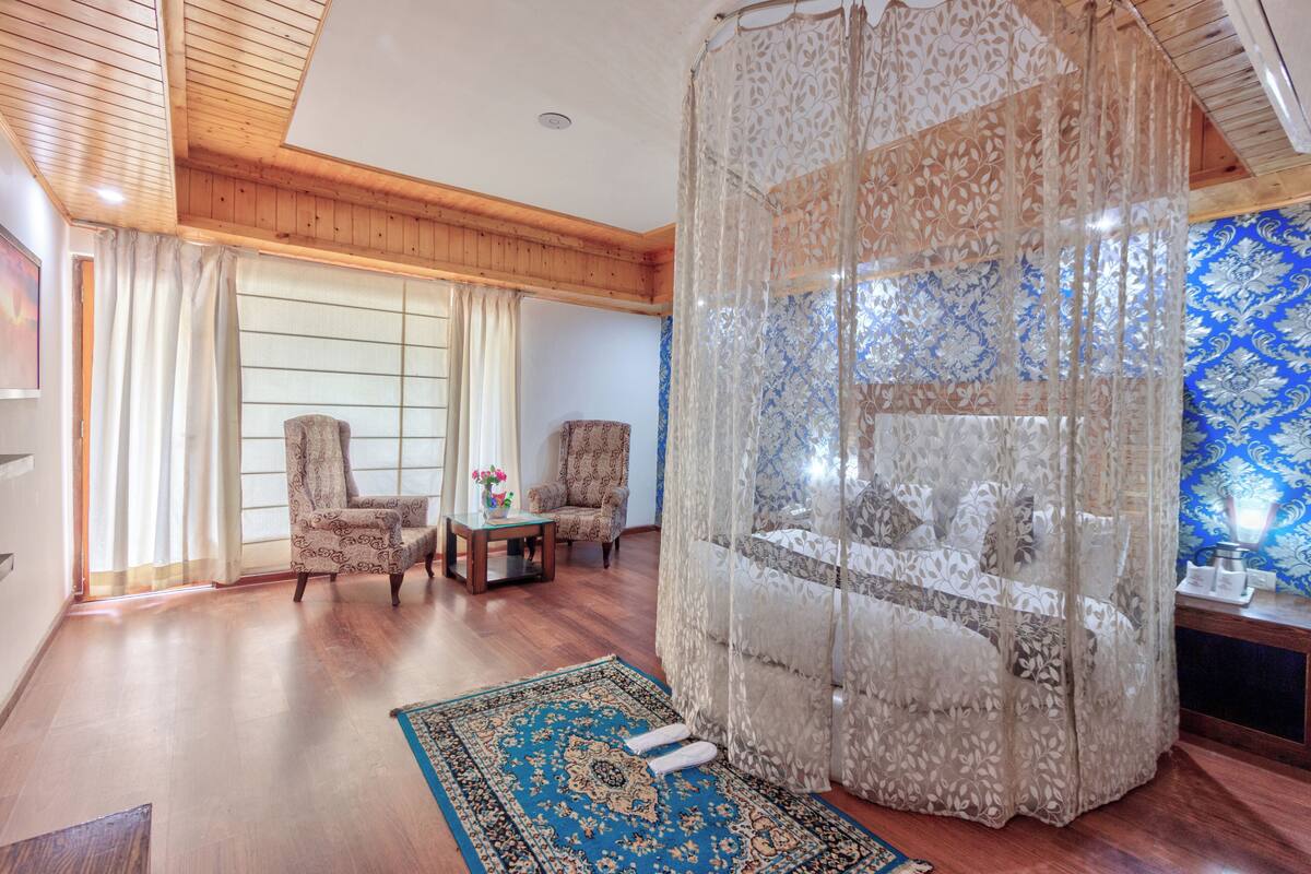 Una habitación con techo de madera, dos sillones estampados, una mesita con un jarrón, una alfombra azul y dorada, y una cama con dosel.