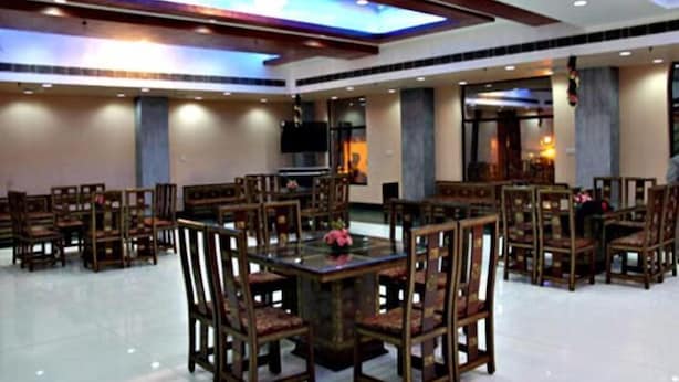 A spacious dining area with multiple wooden tables and chairs, a large flat-screen TV mounted on the ceiling, and a modern ceiling design with recessed lighting. - Sydney Grand Hotel & Resort (New Delhi)