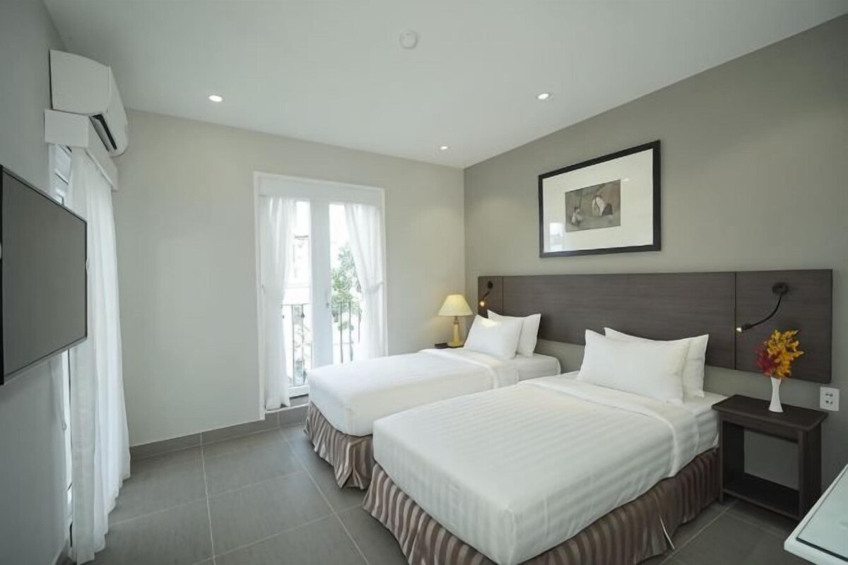 Ein Hotelzimmer mit zwei Betten, einem Fernseher, einem Nachttisch mit Lampe, einem gerahmten Bild an der Wand und einem Fenster mit Vorhängen.