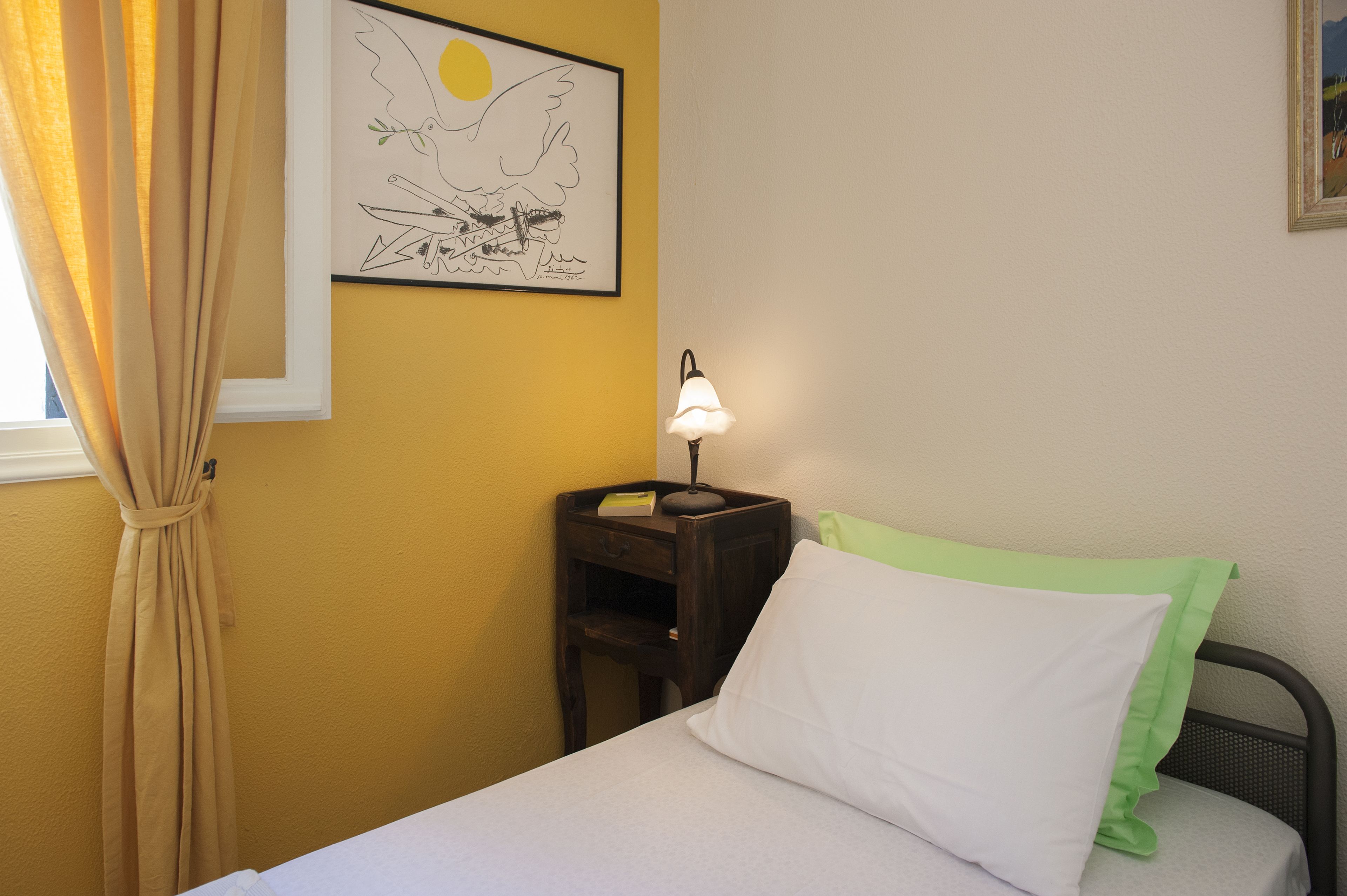 Ein Schlafzimmer mit einem Bett, einem Nachttisch mit Lampe, einer gerahmten Zeichnung an der Wand und einem Fenster mit Vorhängen.