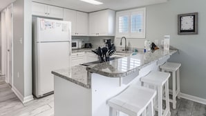 Una cucina moderna con mobili bianchi, frigorifero integrato, microonde, macchina del caffè e lavandino. Sono presenti tre sgabelli da bar bianchi vicino al bancone.