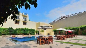 A poolside area with outdoor seating and a building in the background. - Westend Inn (New Delhi)