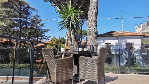 An outdoor dining area with wicker furniture, a table, and chairs, surrounded by a glass fence and trees. - Hotel Bellini (Riccione)