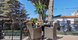 An outdoor dining area with wicker furniture, a table, and chairs, surrounded by a glass fence and trees. - Hotel Bellini (Riccione)