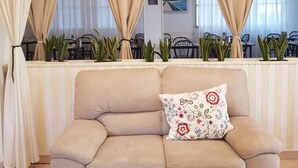 A beige sofa with a floral pillow in a room with beige curtains and a view of a dining area with chairs and plants. - Hotel Bellini (Riccione)
