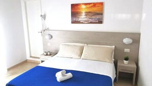 A hotel room with a bed, a painting of a sunset, a bedside table, a nightstand, and an air conditioning unit. - Hotel Bellini (Riccione)