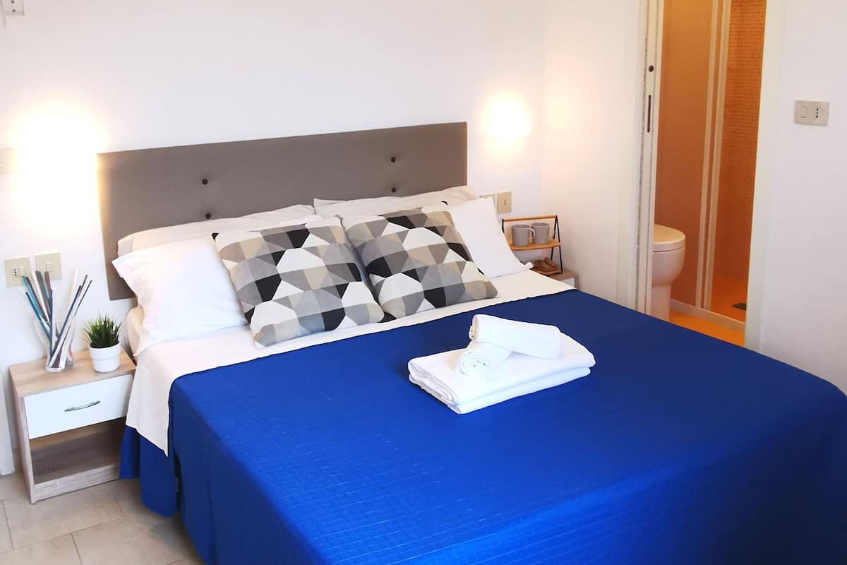 A neatly made bed with a blue bedspread, a pillow with a geometric pattern, and a folded white towel on the bed.
