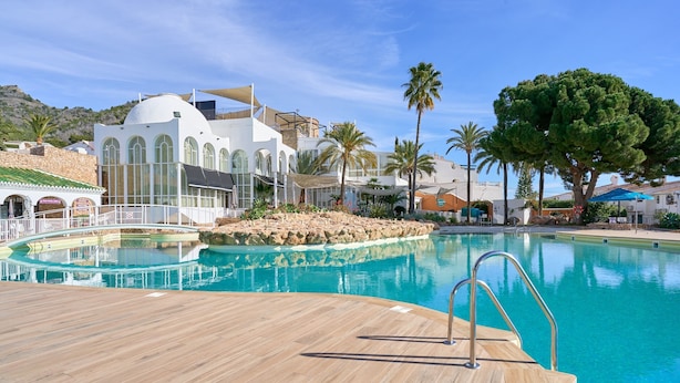 Un área de piscina con cubierta de madera, un edificio blanco con ventanas de arco y palmeras.