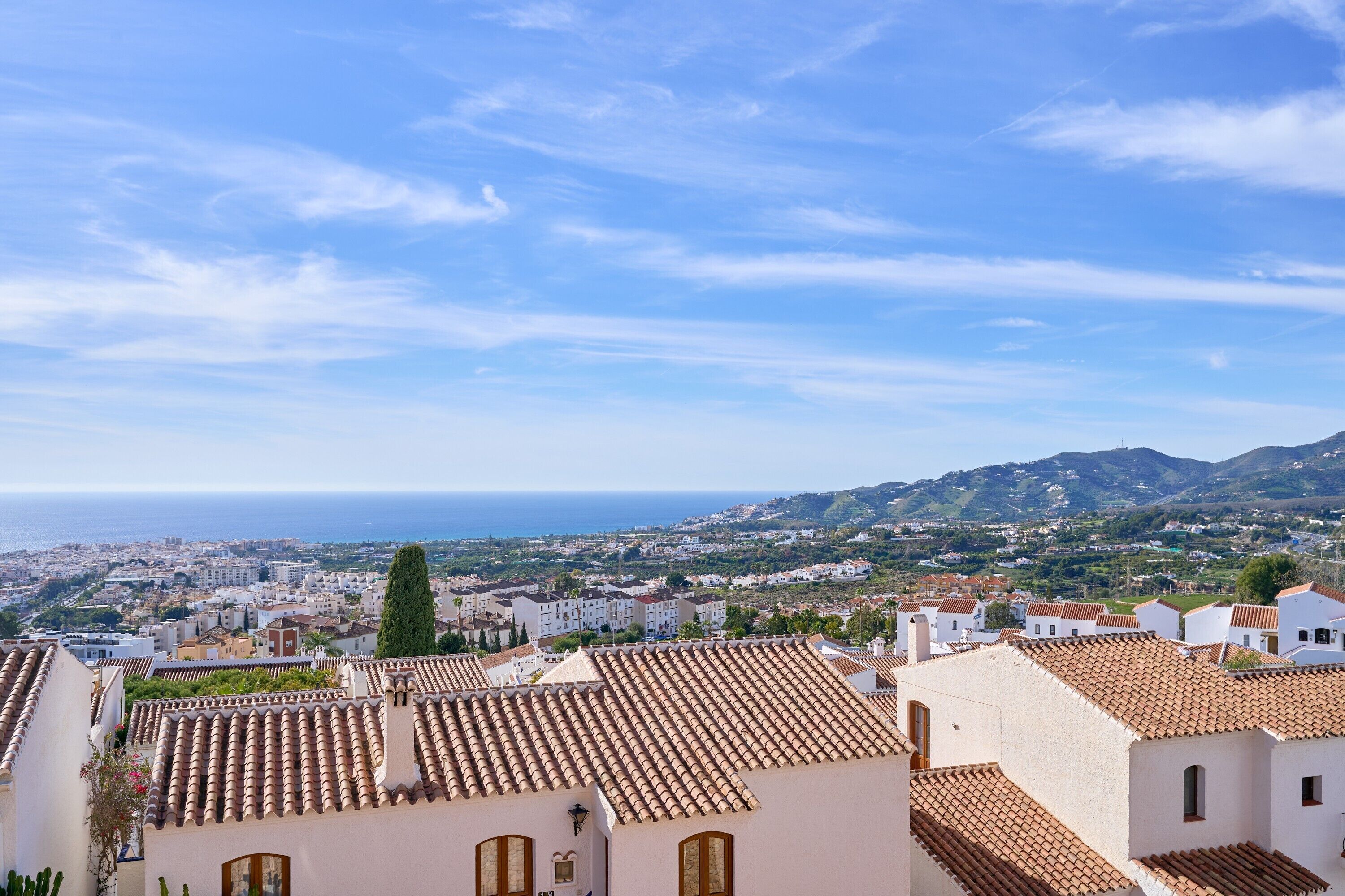 Une ville côtière aux bâtiments aux toits de tuiles en terre cuite, offrant une vue sur la mer et s’étendant sur un paysage vallonné à l’horizon.