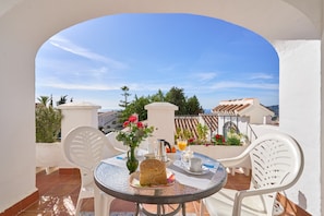 A round table with chairs, a vase with flowers, a bread roll, a coffee pot, and a glass of orange juice on a terrace with a clear view of the sky and buildings.