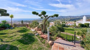 Un jardín ajardinado con vistas a un pueblo costero, un edificio blanco con un campanario y un cielo despejado.